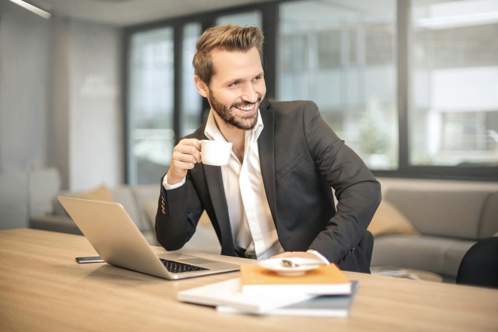 Smiling businessman in formal attire enjoying coffee break at modern office desk with laptop.